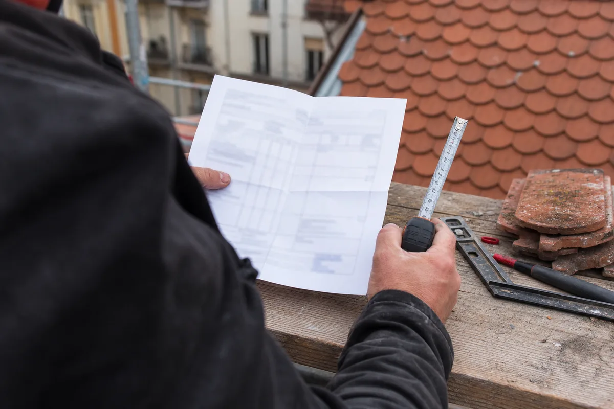Mains d’un couvreur vu de dos consultant un devis de toiture papier avec un mètre ruban sur une table de chantier