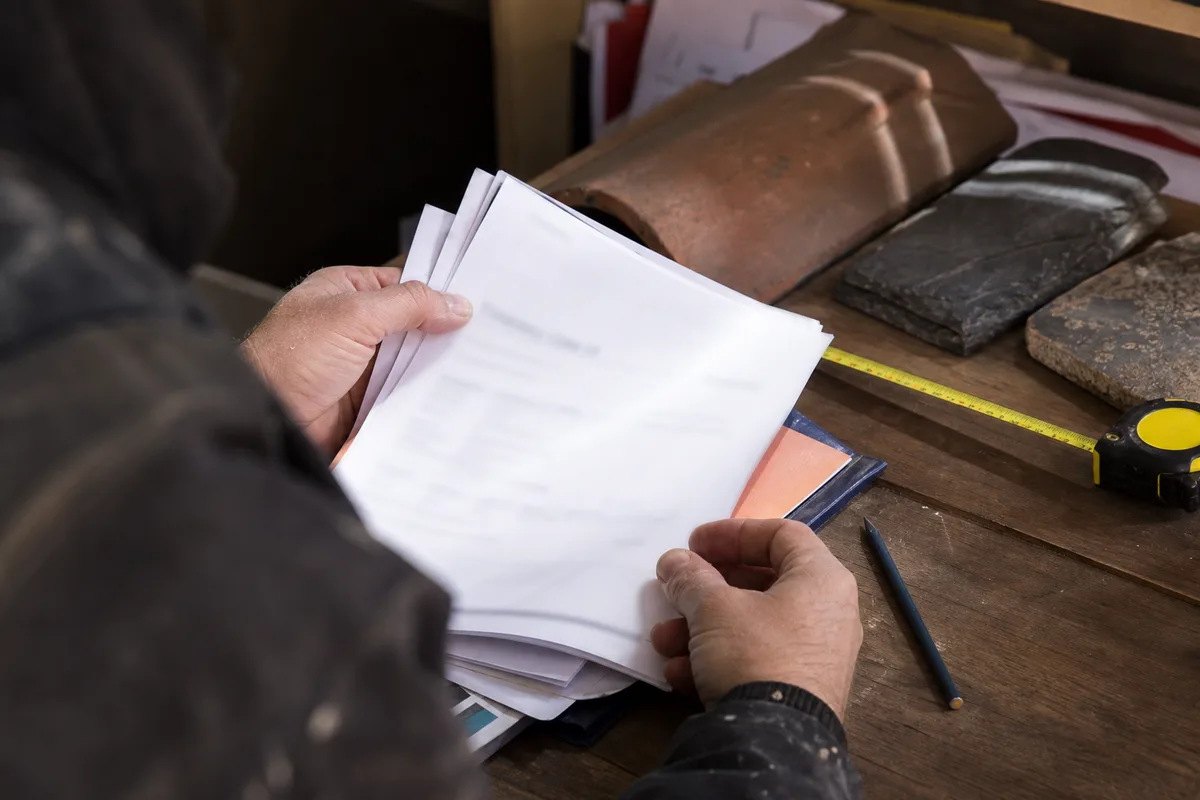 Un couvreur consulte un devis de toiture papier posé sur une table, mètre ruban et échantillons de tuiles à côté