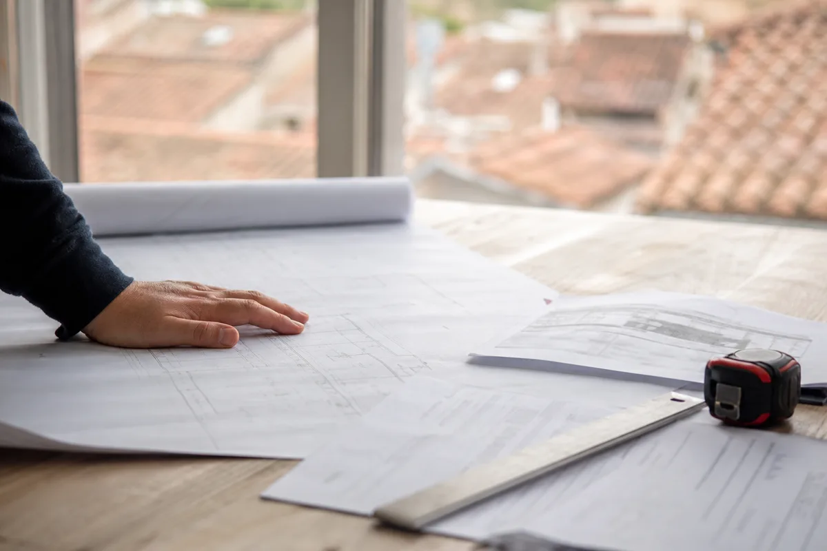 Plan de toiture, mètre ruban et feuilles de devis posés sur une table près d&rsquo;une fenêtre donnant sur les toits
